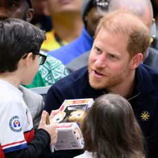 Prince Harry wearing a blue sweater talking to a young boy who is holding a Funko Pop Prince harry doll 