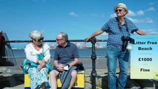 3 older people by a beach railing, with Martin Parr in a straw hat and sunglasses holding a "Litter Free Beach" sign to the right, and a couple seated to the left