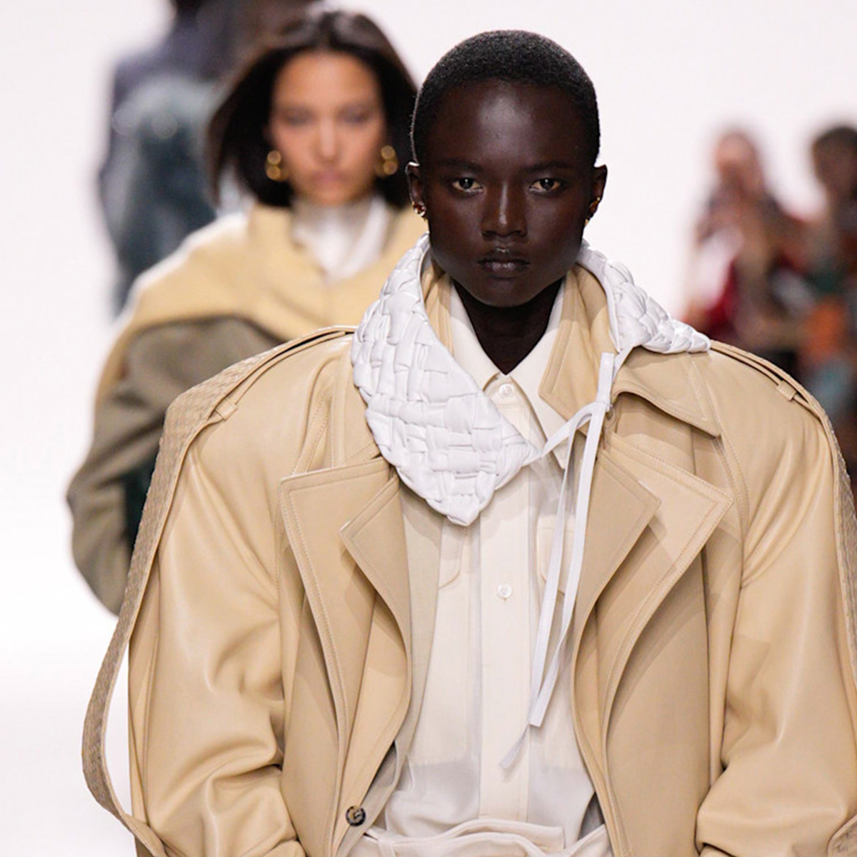 A model at the Bottega Veneta S/S 26 show wears a tan oversized leather trench coat with a white collar. 