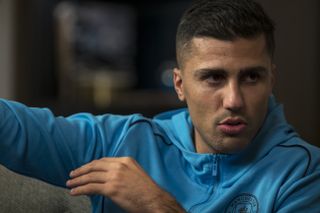 Close up of Manchester City midfielder Rodri in a blue Manchester City hoodie guesting to his left as his is interviewed