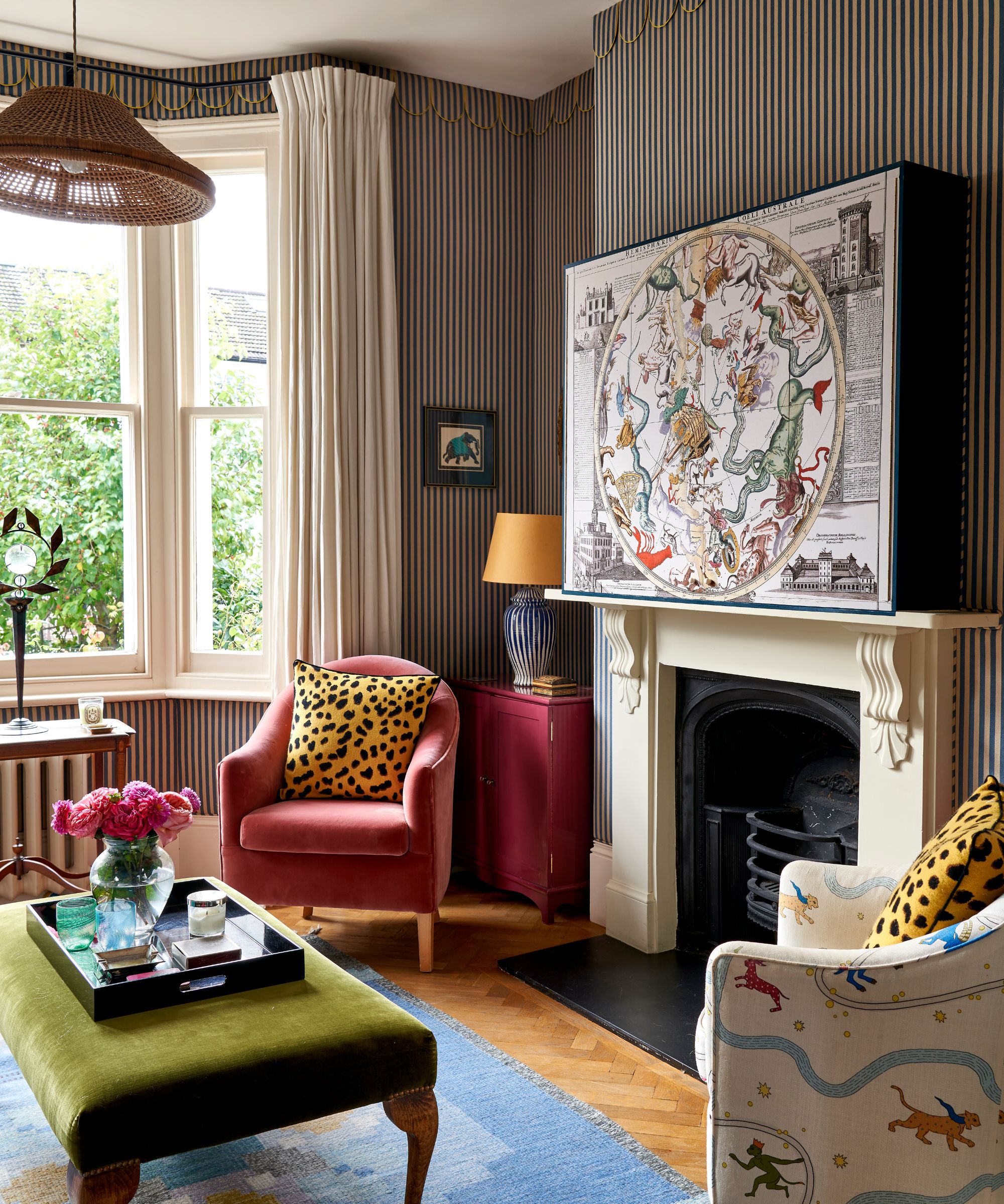 a striped living room with traditional fireplace, large artwork box hiding a TV, a small pink armchair with a leopard print pillow and a series of eclectic side tables in the bay window and alcove