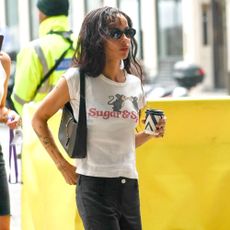 Zoe Kravitz walks outside holding a coffee wearing a white graphic t-shirt, black trousers and black heeled peep-toe shoes.