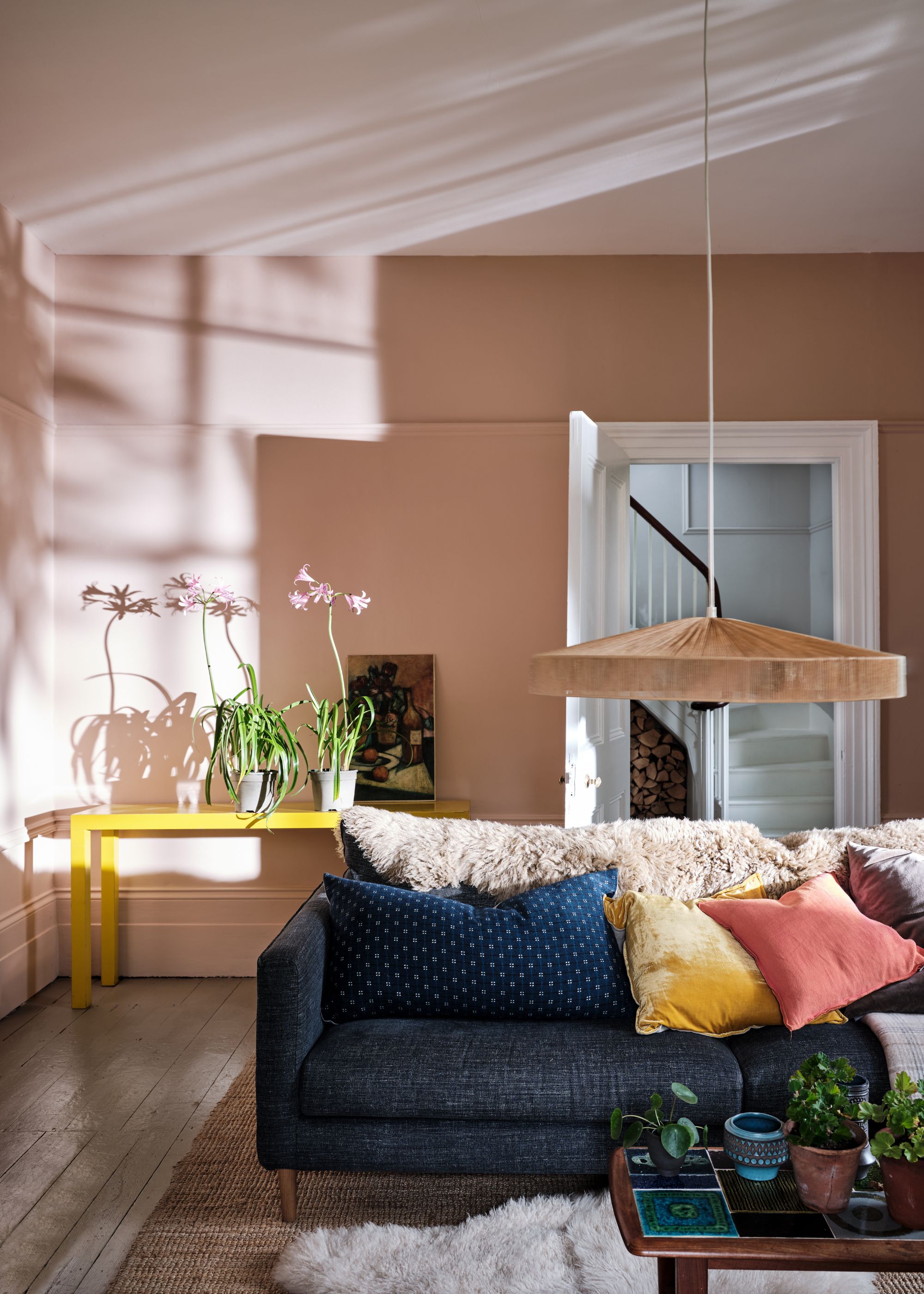 Living room with plaster pink walls, navy blue sofa with pink, blue, and yellow cushions, white ceiling and trim, and yellow side table in the corner