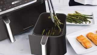 A Ninja air fryer sitting next to a basket as someone removes asparagus from it