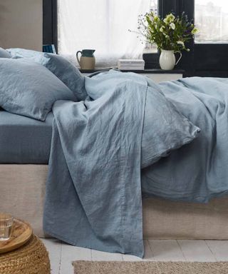 Side view of dusk blue bed sheets on a bed against a white wall and windows.