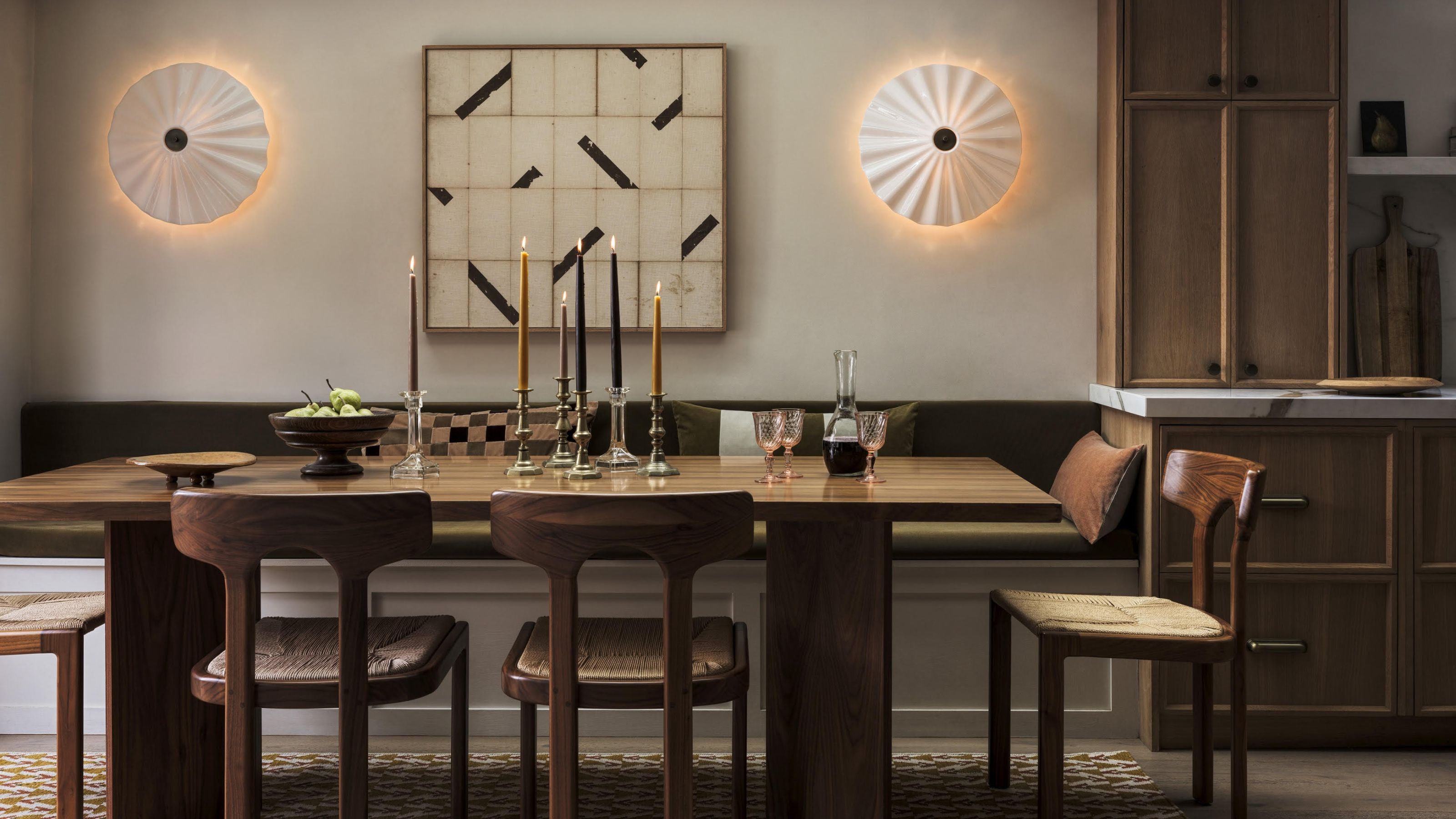 A dining room with large wall lights between a painting in the middle of the room. On the dining table is a group of candles as well as a bowl of fruit and glassware.