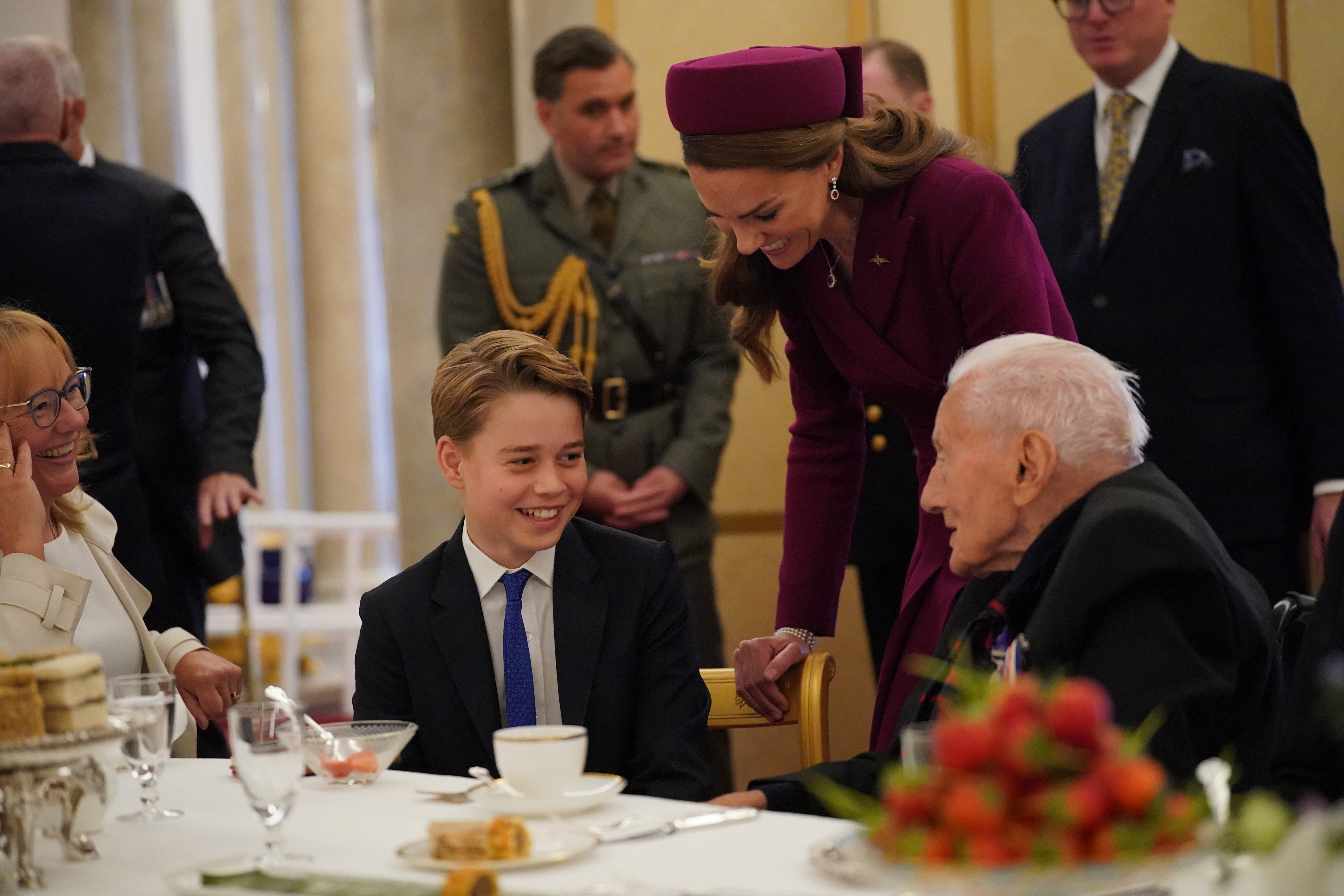 Prince George sitting at a table smiling at a WWII veteran with Princess Kate standing behind him