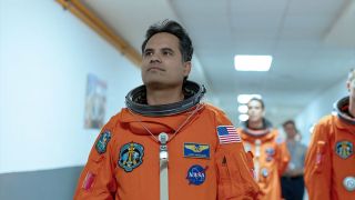 an astronaut in an orange space suit smiles while walking down a hallway