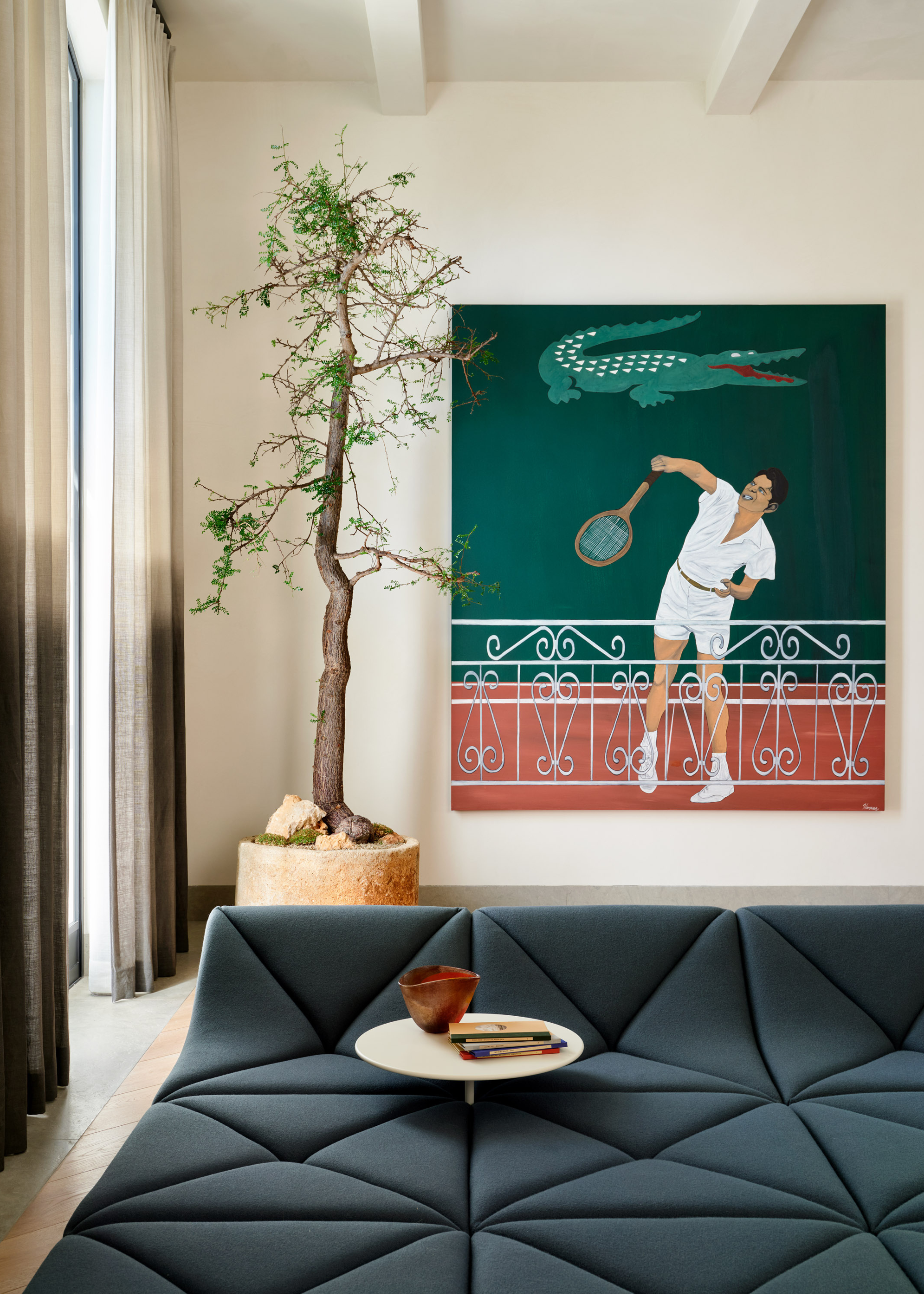 A white living room with a tennis-themed Lacoste painting beside a potted indoor tree and a floor sofa with a floating side table