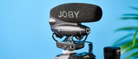 a black shotgun microphone by joby called the wavo pro is photographed by itself against a blue background, showing its muff, dead cat, attachment foot, and connection ports