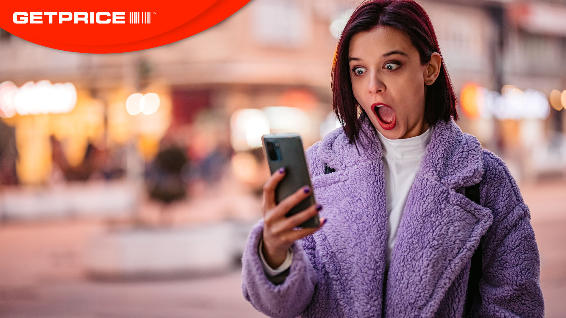 Woman looking at her phone with a shocked/excited face, wearing purple jacket with Getprice logo on top left corner