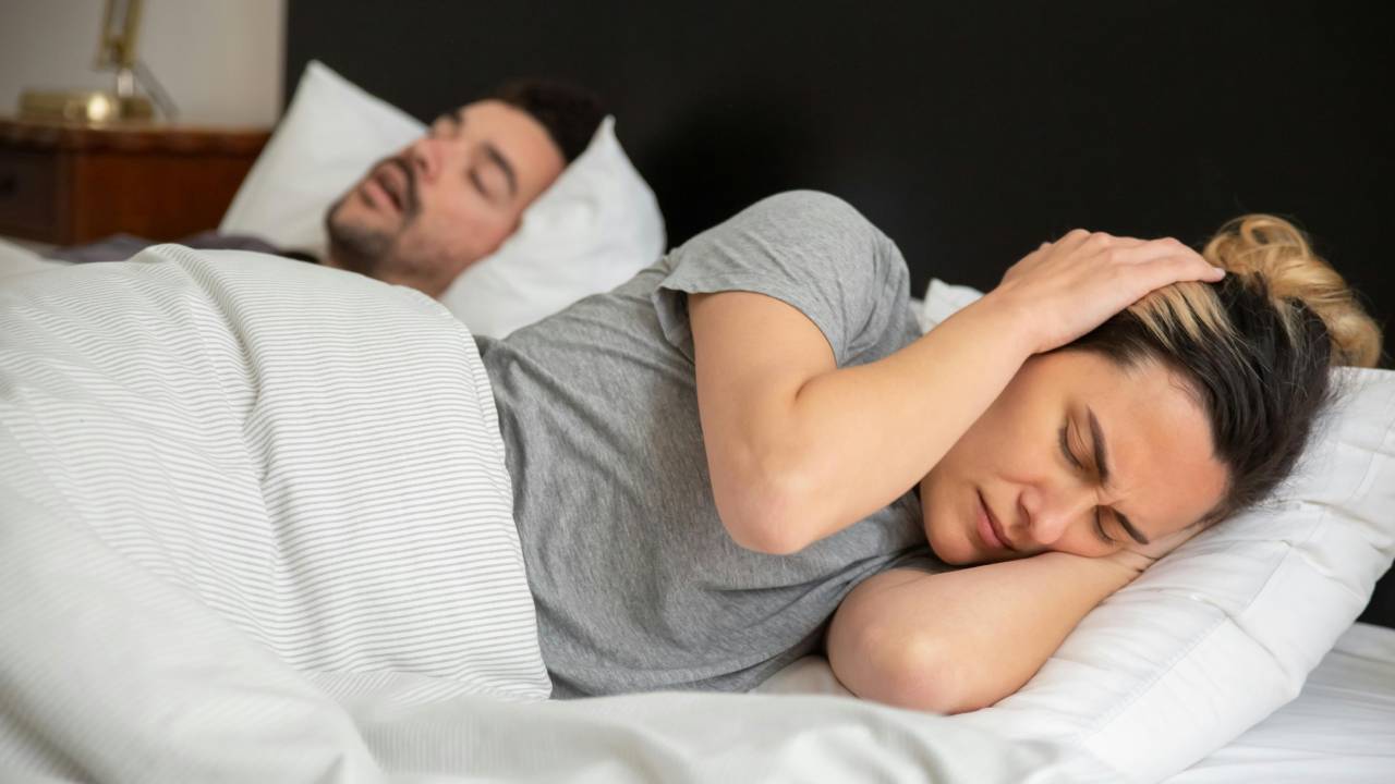 A man lying on his back snoring while a woman covers her ears