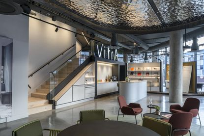 Inside a spacious VitrA showroom two tables and chairs immediately in front, an island area behind featuring a large illuminated VitrA sign, and a stairwell that leads to an upper level. 