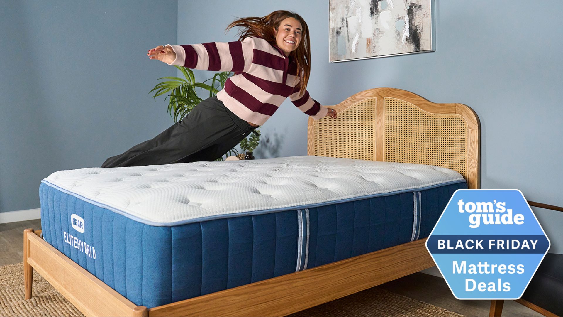 Our mattress tester jumping on the Bear Elite Hybrid mattress on a wooden bedframe with a rattan headboard in our sleep studio with blue walls and a bedside plant