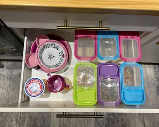 An open kitchen drawer with six colorful cereal bins with cereal visible through the window in the top. On the left of the drawer is a stack of colorful children's bowls