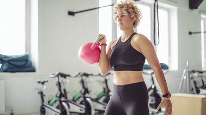 A woman stands in a gym holding a kettlebell at shoulder height in her right hand; her other hand hangs at her side. Behind her we see a row of exercise bikes and large windows.