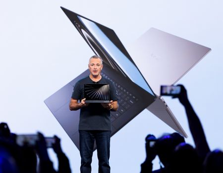 Man presenting a sleek laptop on stage, with a large image of the device projected behind him. Audience members capture the moment on phones.