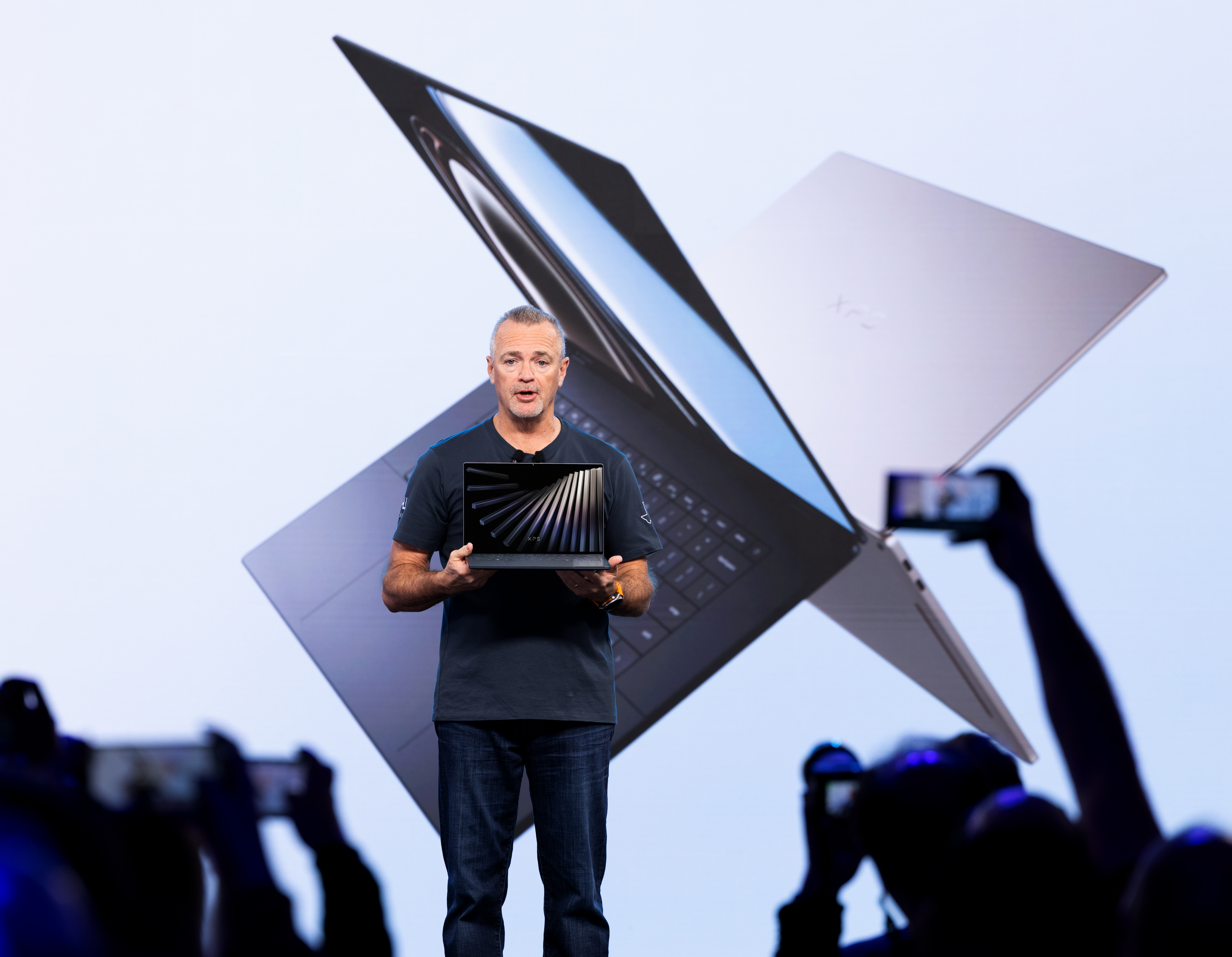 Man presenting a sleek laptop on stage, with a large image of the device projected behind him. Audience members capture the moment on phones.