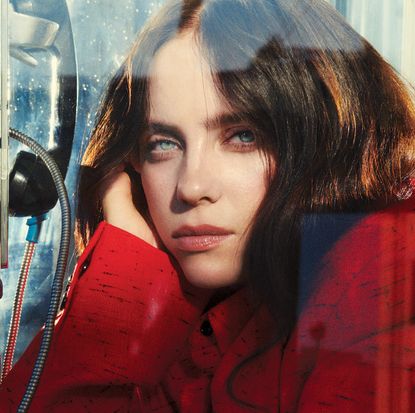 Billie Eilish wearing a red coat posing with her hand on her cheek inside of a phone booth