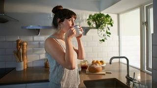 A woman standing at window with mug of tea in kitchen with morning light coming in.