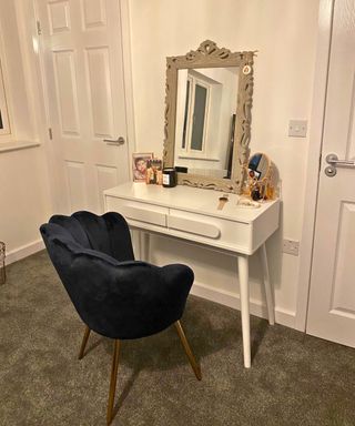 Navy blue velvet clam chair with gold legs in front of a white vanity table with gray ornate mirror in white bedroom