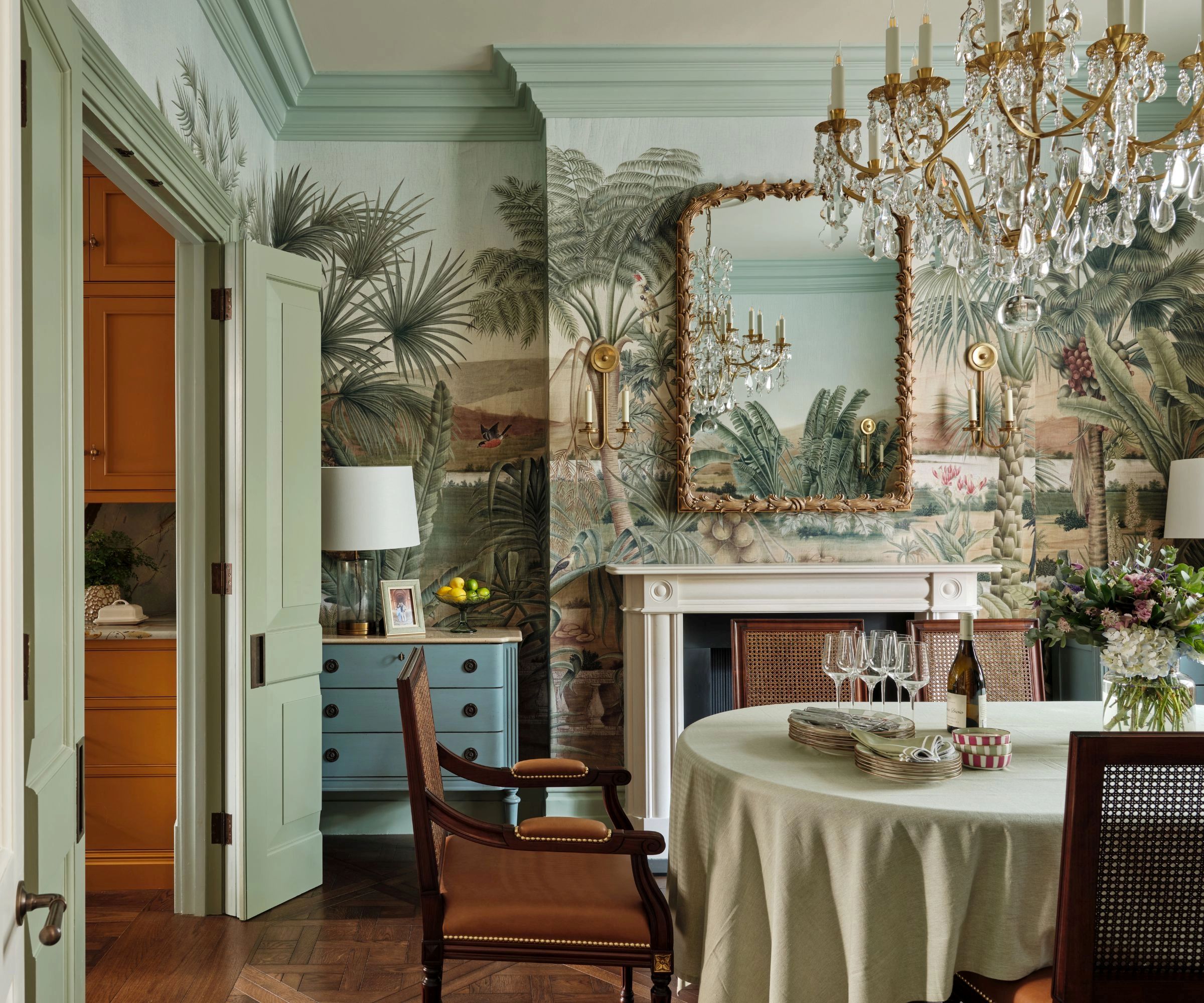 maximalist dining room with mural wallpaper, soft green painted trims, a large crystal chandelier with an antique table and chairs
