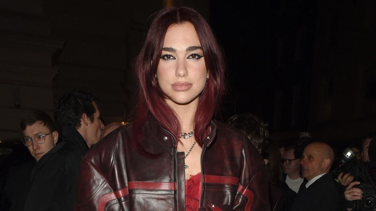 Dua Lipa wearing a brown and red leather coat over a red negligee lingerie dress with tights and red heels