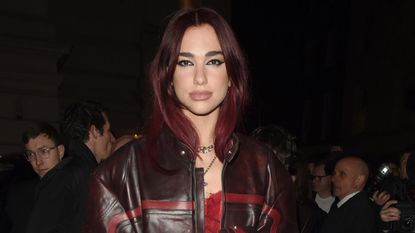 Dua Lipa wearing a brown and red leather coat over a red teddy lingerie dress with tights and red heels
