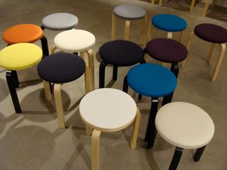 Multiple round three-legged stools in different colours, photographed on a grey surface