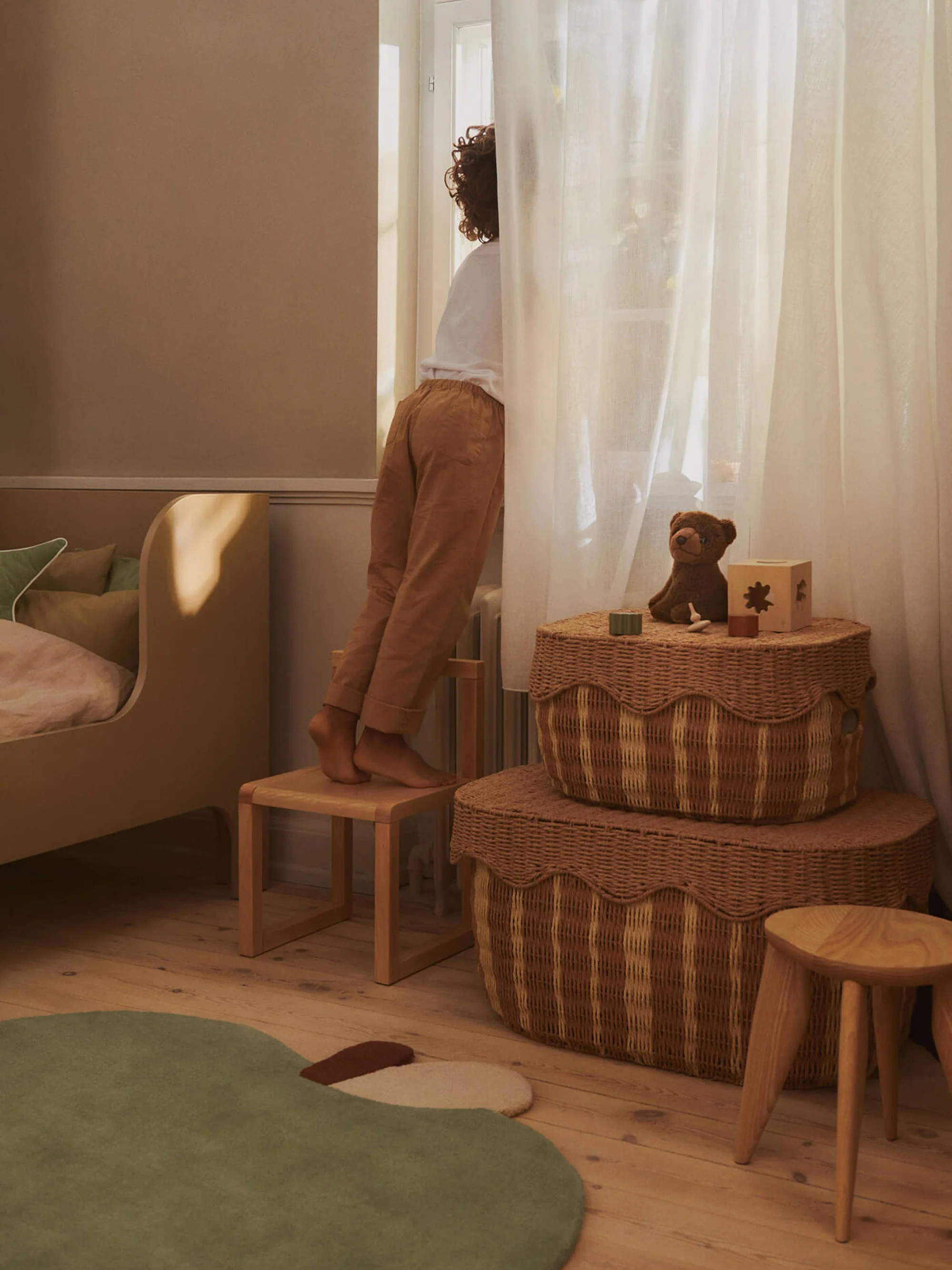 Child standing on a wooden stool beside a softly lit window in a warm, neutral-toned bedroom with wicker storage baskets, sheer curtains, and natural wood furniture.
