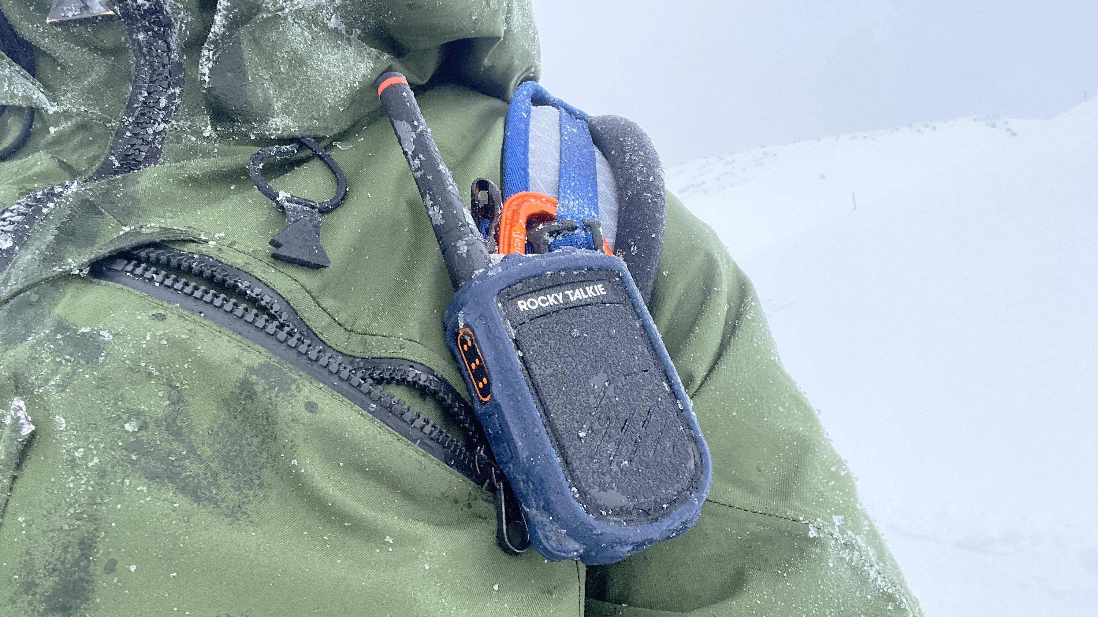 Close up dari walkie talkie radio talkie rocky mountain yang melekat pada tali ransel biru di atas jaket ski hijau yang ditutupi es dengan salju di kejauhan