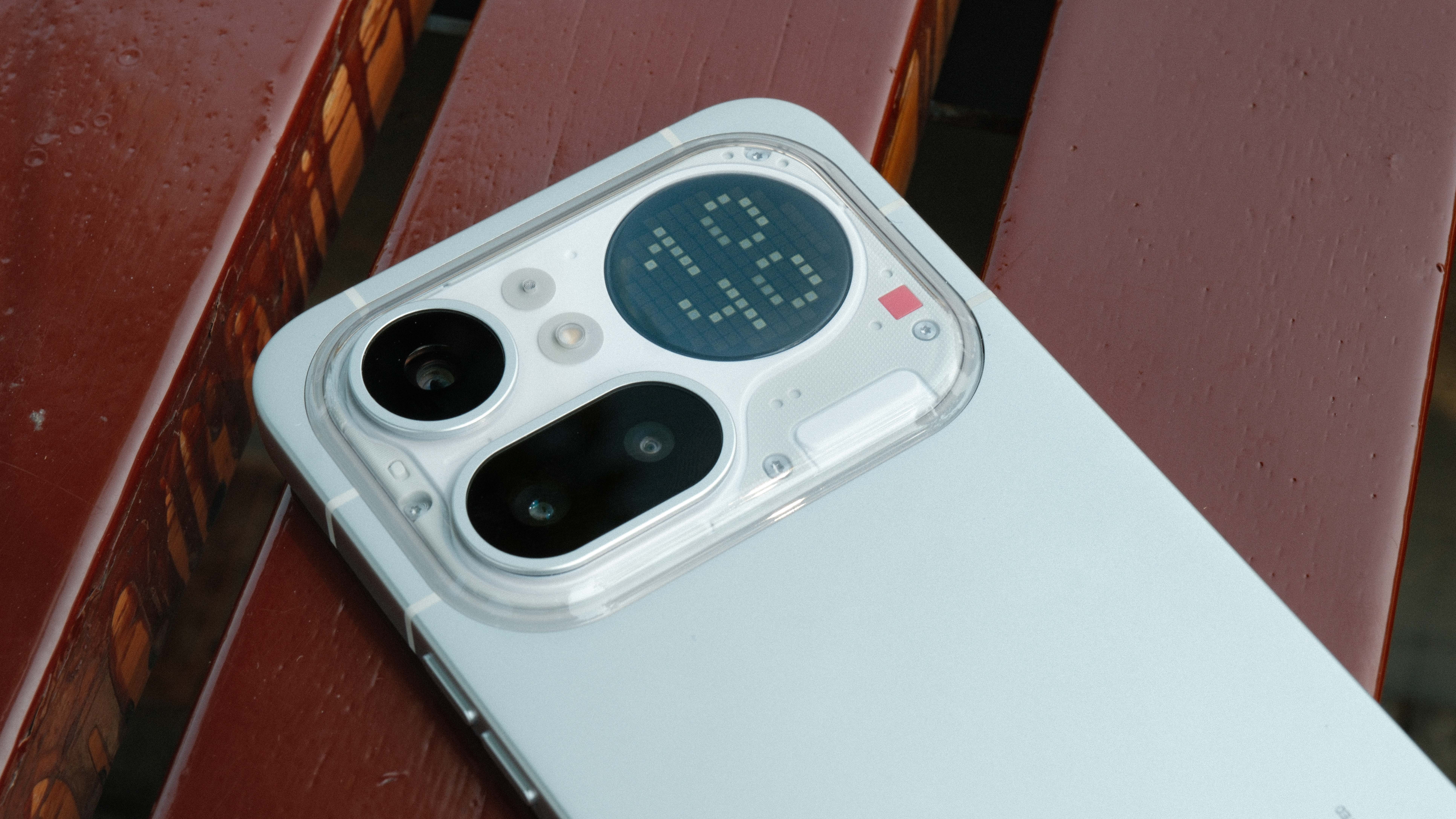 A close-up, high-angle shot of the upper back of a light gray smartphone resting on a dark wood slatted surface. The transparent camera island features three distinct lens housings and a circular dot-matrix display showing the number 43.