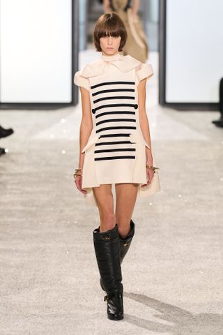 A model at the Sacai spring 2025 show in look 003 wearing a striped dress and black knee high boots