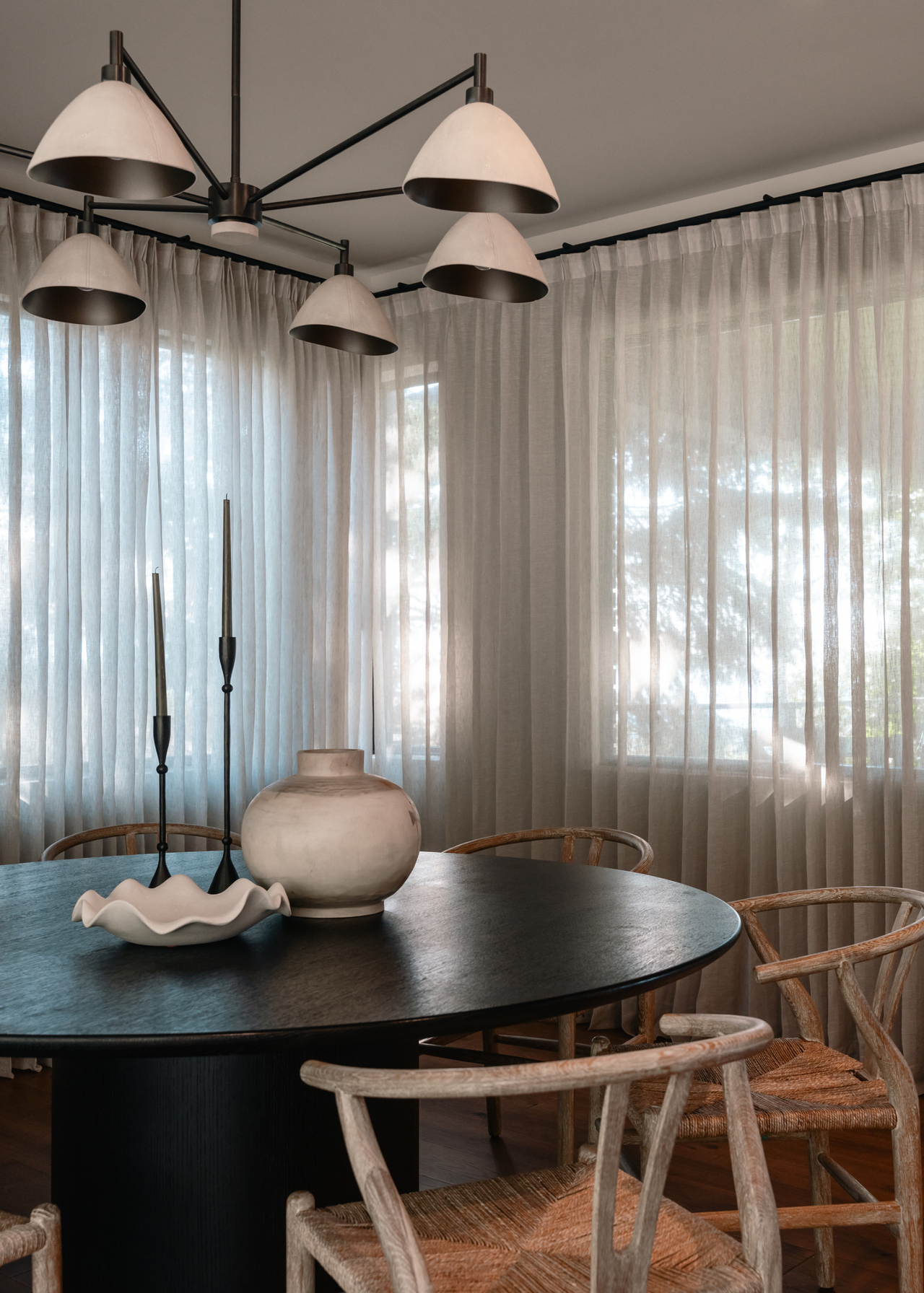 A dining room with sheer curtains dappling light, a round table, and wood chairs. Also pictured are ceramic vases and black candlesticks and holders