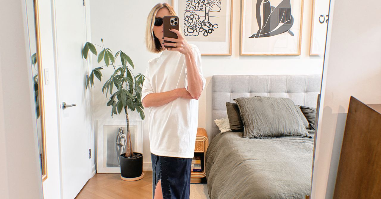 Natalie Cantell taking a mirror selfie in her NYC apartment wearing an oversize white Tibi t-shirt, denim Tibi skirt, Celine eyewear, and brown Havaianas flip-flops.