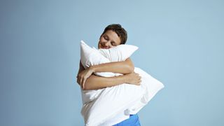 A woman with short dark hair hugs a white bed pillow while stood against a bright blue wall