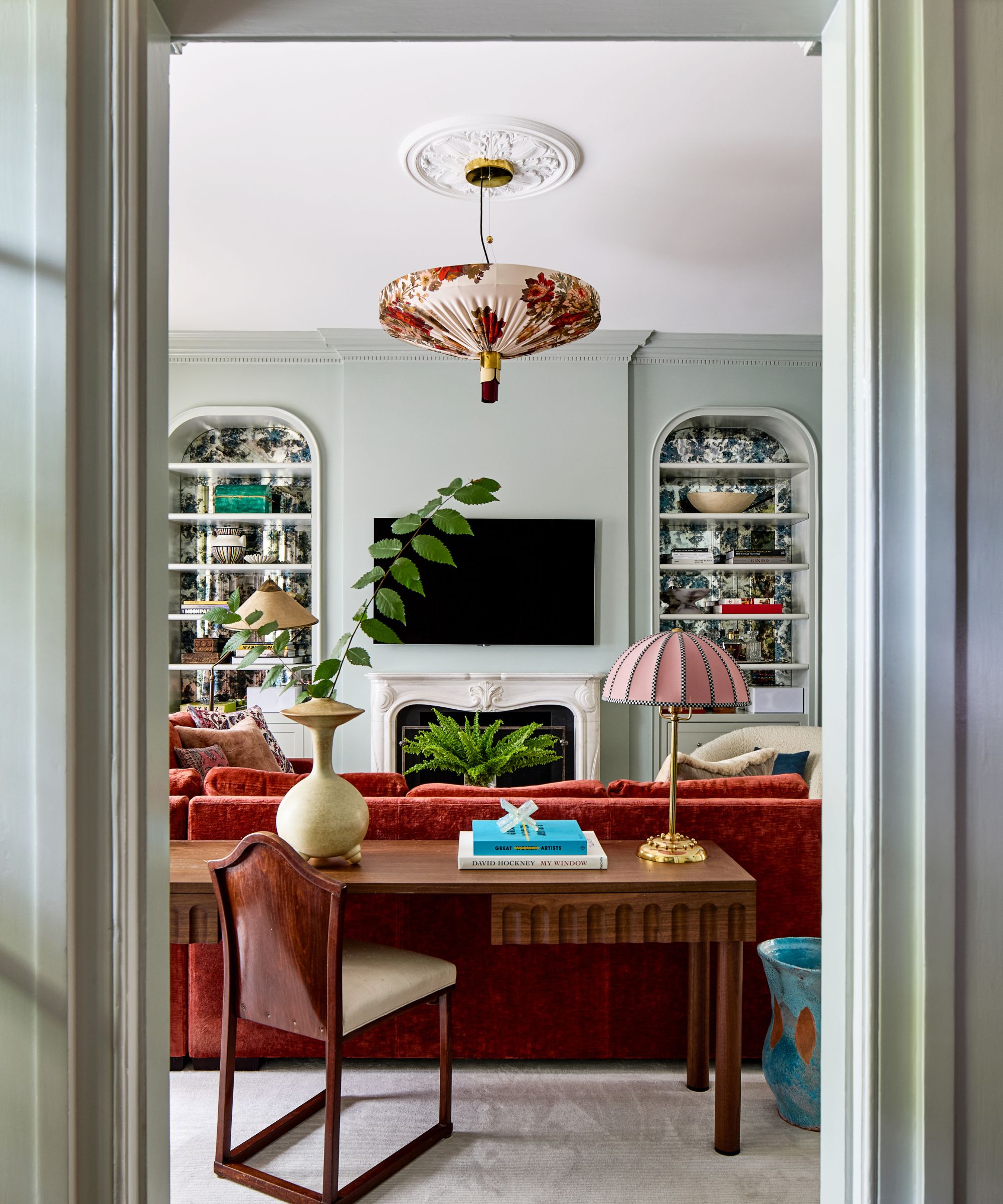 Living room with sage green walls, fireplace and TV above it, red sofa, and a wooden desk with a pink table lamp, vase and books on top of it behind the sofa