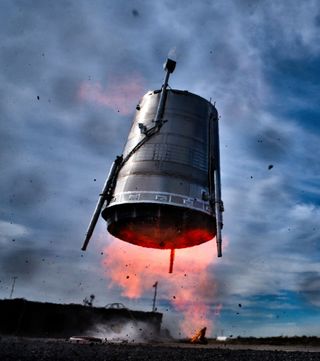 a cylindrical silver rocket stage flies low to the ground, with orange flames erupting from its base.