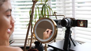 A person in front of a makeup mirror, filming themself applying makeup on a Fujifilm X-M5 camera