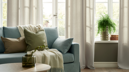 IKEA GINSTMOTT Curtains in Beige against white-framed windows with a blue sofa in front with cushions and a throw, as well as a coffee table with a green vase with a singular stem of foliage inside