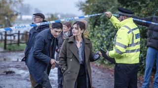 Archie Renaux in a suit as Hitch and Jenna Coleman in a dark coat as Ember walk under some police tape being held up by an officer in a hi-vis jacket in The Jetty