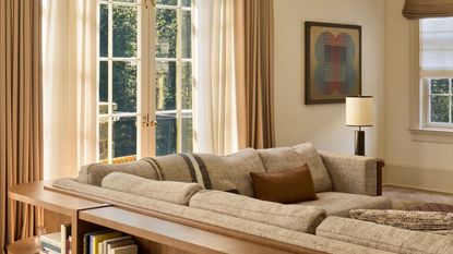 A modern neutral living room with cream walls, French doors with layered neutral curtains, a large taupe corner sofa, with a wooden console table running along the back, with styled books. 