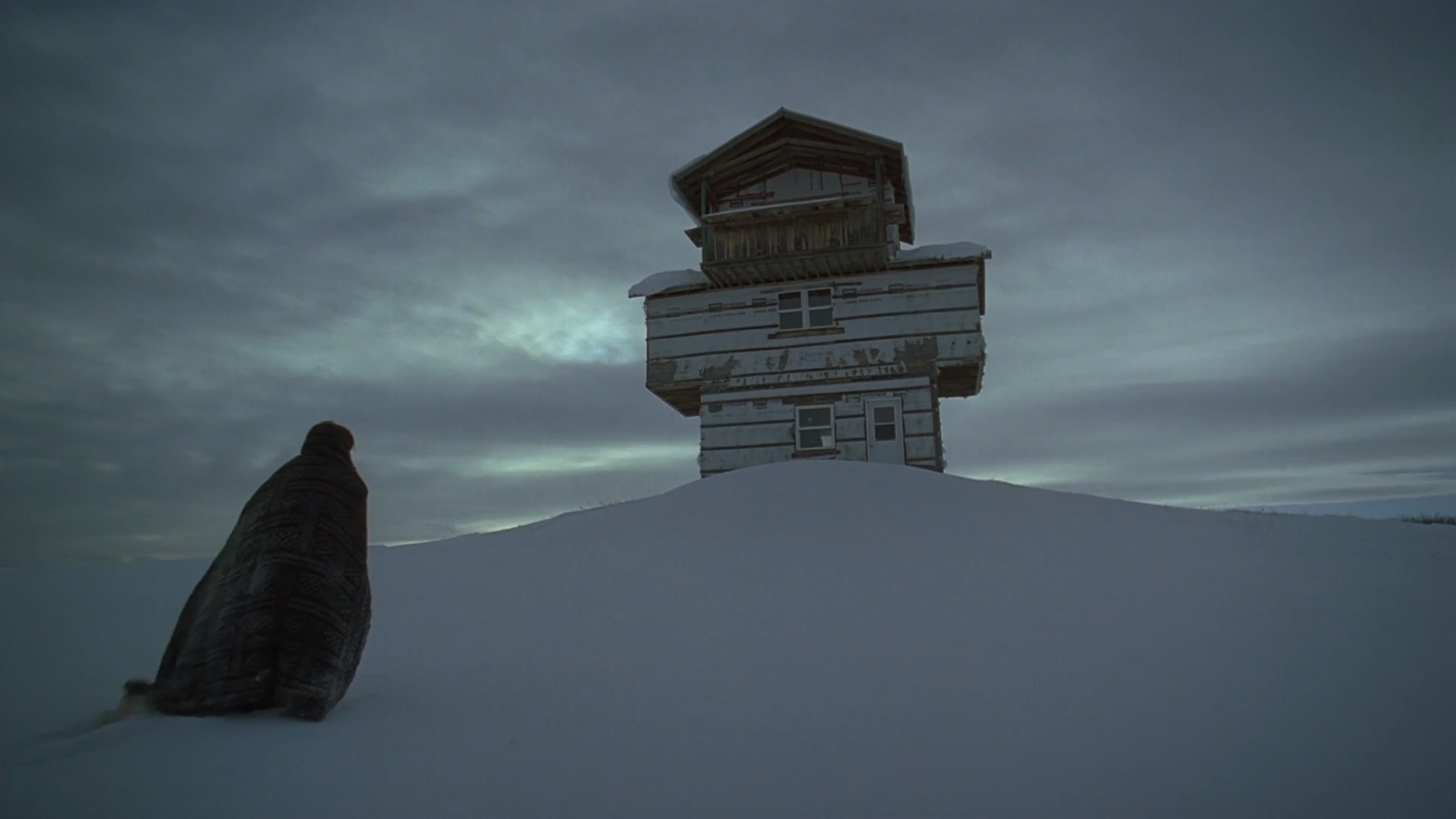 A cloaked figure in front of a building on a snowy night in The Lodge, one of the best scary movies on Hulu