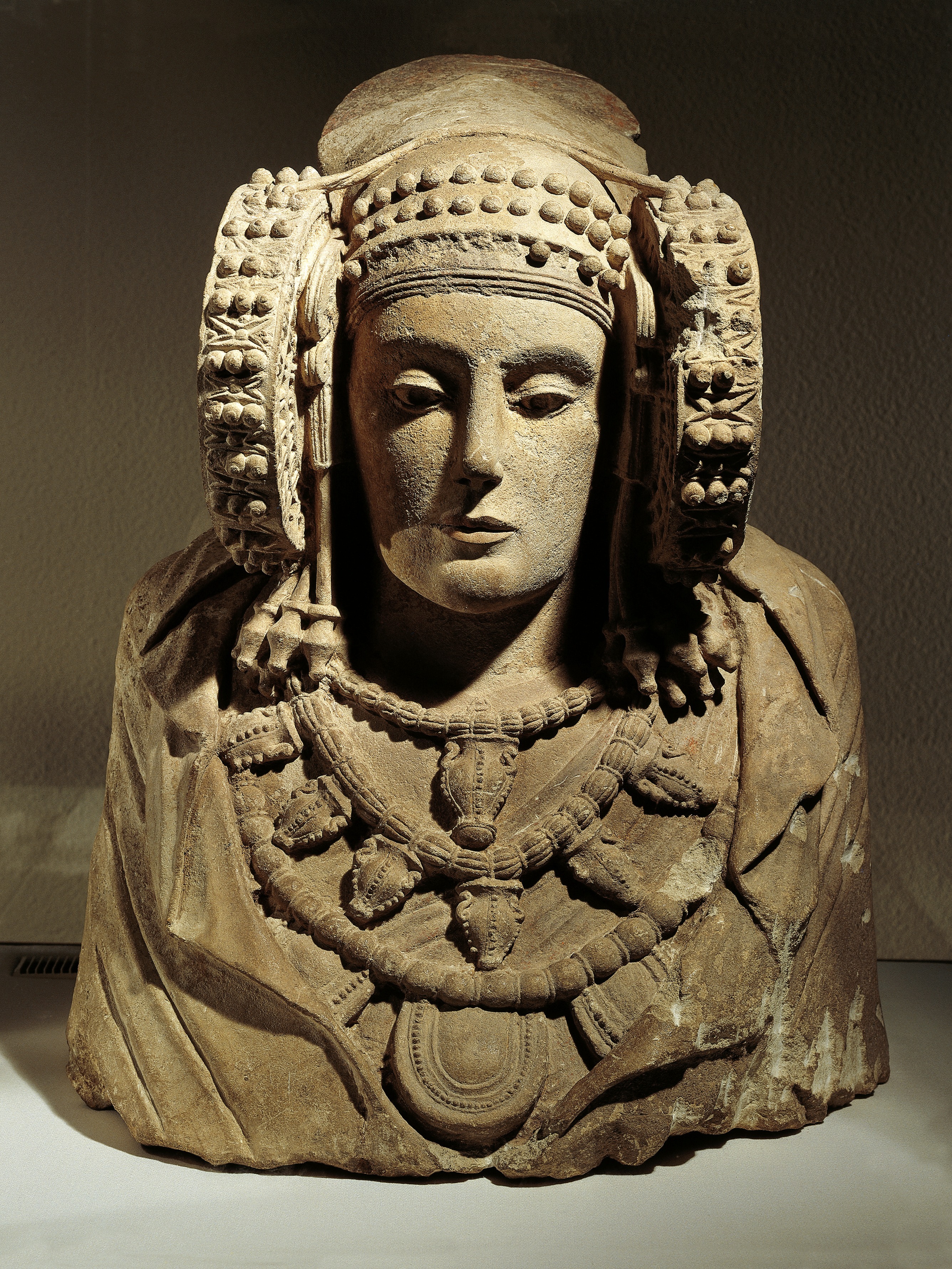 a limestone bust of a woman wearing a cloak, jewelry and large circular earrings