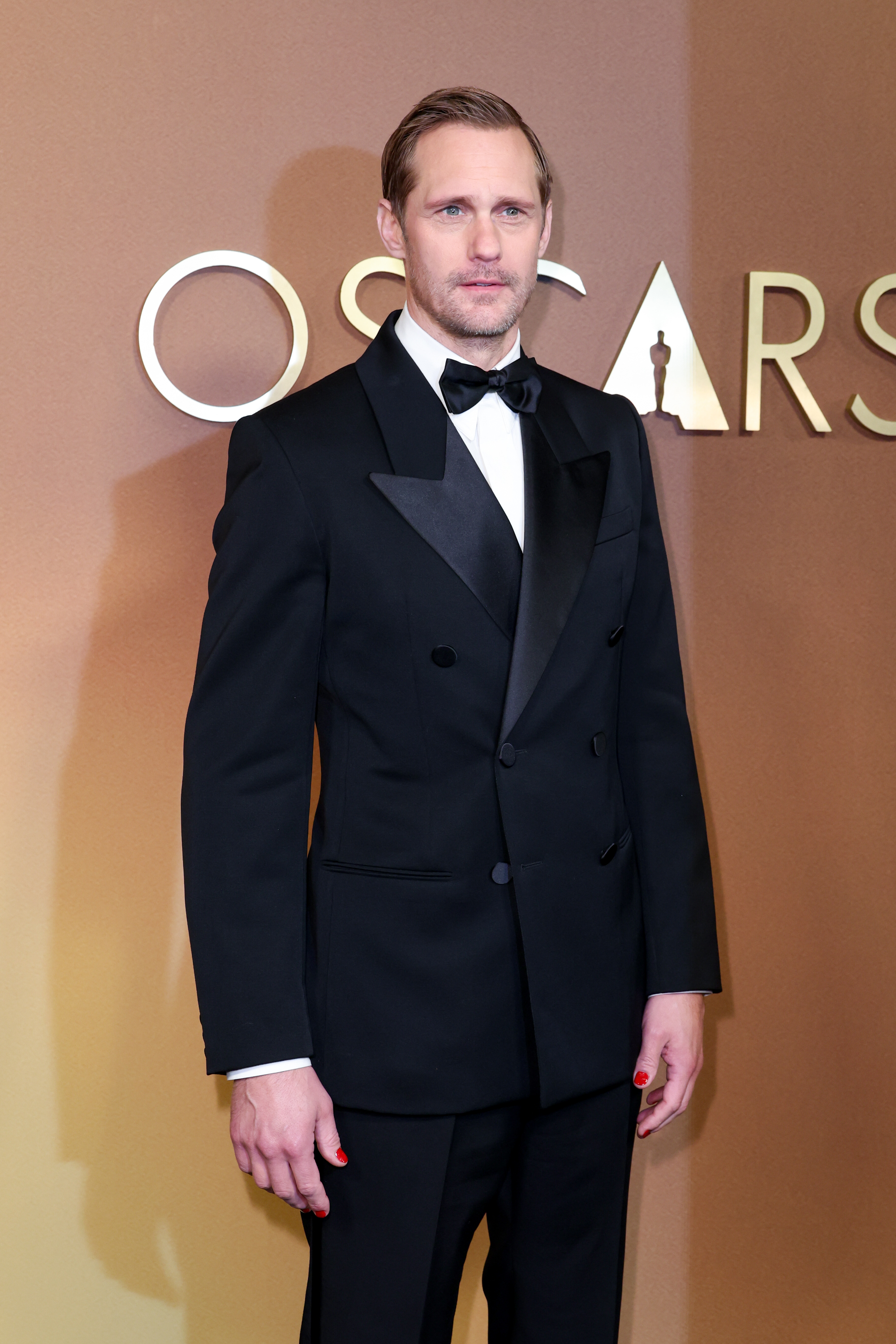 a white man wearing a tuxedo and posing in front of an amber background at a red carpet event