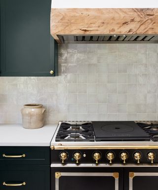A kitchen with white weathered zellige tiles, a black and brass range, green cabinets and oak hood