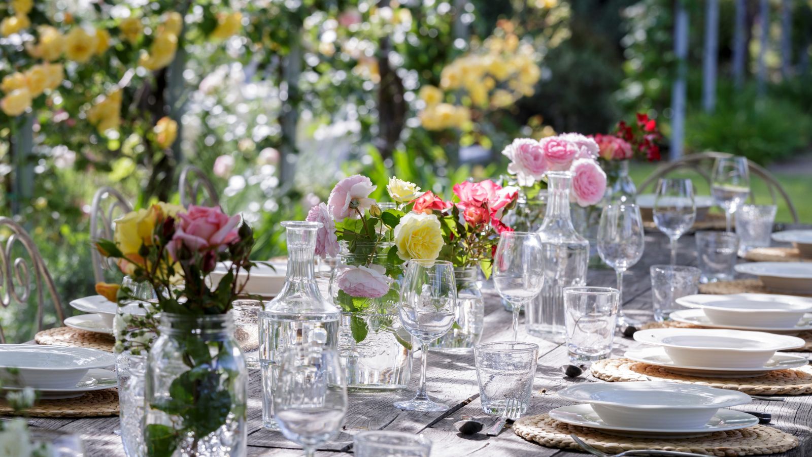A summery tablescape in a garden with lots of summer flowers such as pink and yellow roses in glass vessels in the center of the table and yellow roses growing in the background