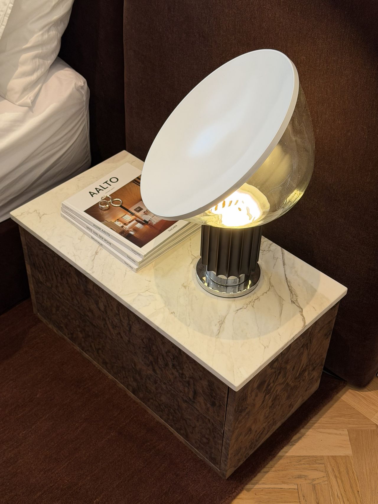 Image of a burl wood nightstand with a white marble top in front of a dark red extended headboard with a matching rug underneath.