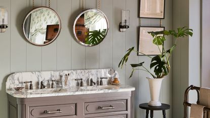 Sage green paneled bathroom with cream and gray veined marble double vanity. There are two round mirrors hanging from chains above each sink and vintage wall lamps