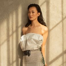 Actress Mari Yamamoto is photographed against a white textured wall. She is wearing a strapless dress with a painted print mimicking a belt. 